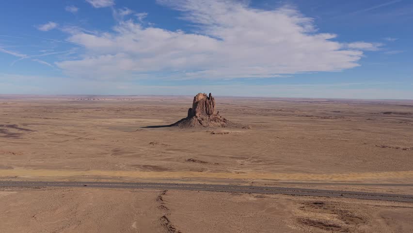 Drone flight moves toward a lone desert butte and highway road trip featuring a car driving through a vast arid landscape with rugged geological formations under a bright blue sky.
