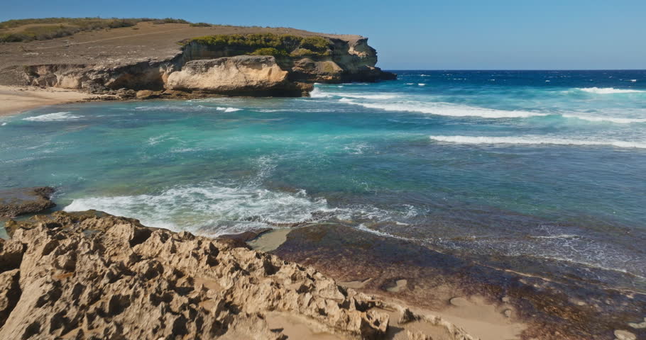 Lombok, Pantai Penyisok rugged coastal cliffs with eroded sandstone rock meet vibrant blue ocean, waves crashing into white foam. Aerial drone flight over remote, untouched tropical shoreline beauty
