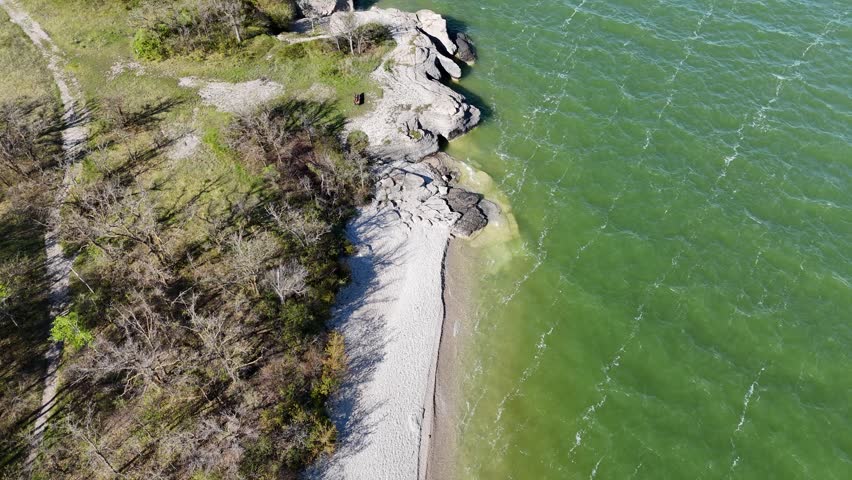 Drone flying overhead along the shoreline at Steep Rock Manitoba showing limestone cliffs, beach, green water of Lake Manitoba, and trails through trees on a summer day.