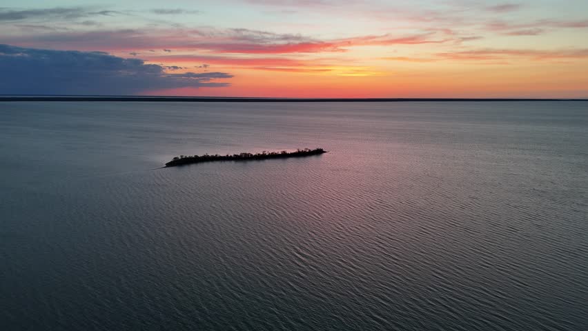 Drone flying toward Goat Island near Steep Rock Manitoba keeping the island centered, showing clear blue water, sunset sky with clouds, and red orange light over Lake Manitoba.