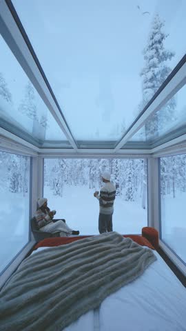 Enjoying a warm drink while surrounded by mesmerizing snowy landscapes, two friends relax in a modern glass cabin in Akaslompolo, Lapland, Finland, embracing the winter magic.