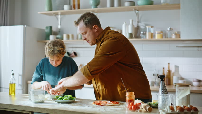 Dad boy making pizza adding toppings together home kitchen closeup. Happy father son preparing tasty Italian cuisine dish enjoying culinary hobby. Caring man sprinkling dough with mozzarella cheese.