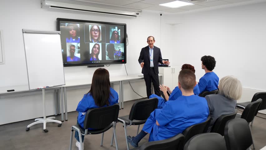 Group of medical students in blue scrubs attending a hybrid conference with a professor presenting in a modern classroom, connecting with remote colleagues via a large interactive digital screen