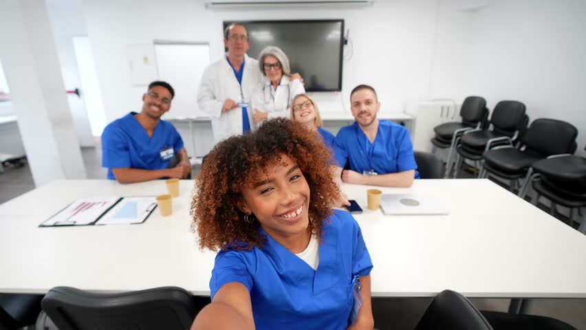 Cheerful group of diverse doctors and nurses taking a funny selfie with a smartphone, smiling and posing together during a break in a modern hospital meeting room after a medical congress