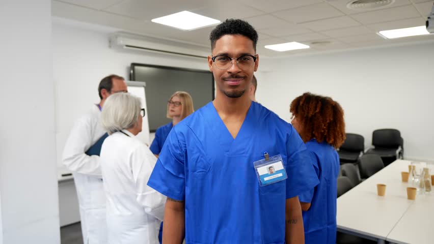 Portrait of a young african american male nurse smiling at the camera. Wearing blue scrubs and an id badge. With his multicultural medical team discussing in the background of a hospital meeting room