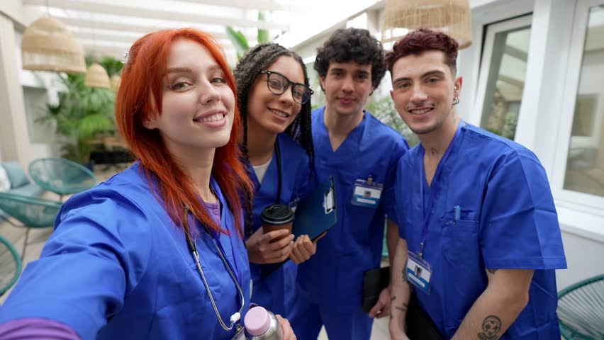 Cheerful group of diverse young medical students in blue scrubs taking a selfie together during their break, smiling and laughing in a modern hospital common area
