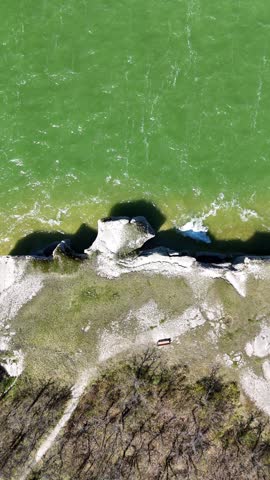 Vertical drone shot of limestone cliffs at Steep Rock Manitoba showing a couple of seagulls flying under the camera, waves on Lake Manitoba, shoreline, trees, and forest in summer daylight.