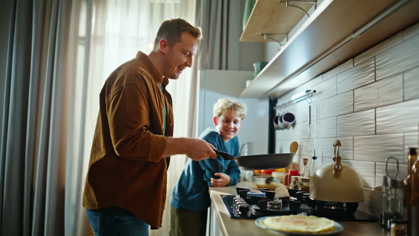 Dad child preparing pancakes together enjoying cozy domestic morning home closeup. Smiling father throwing up turning over tasty breakfast at frying pan. Happy son watching daddy learning cooking.