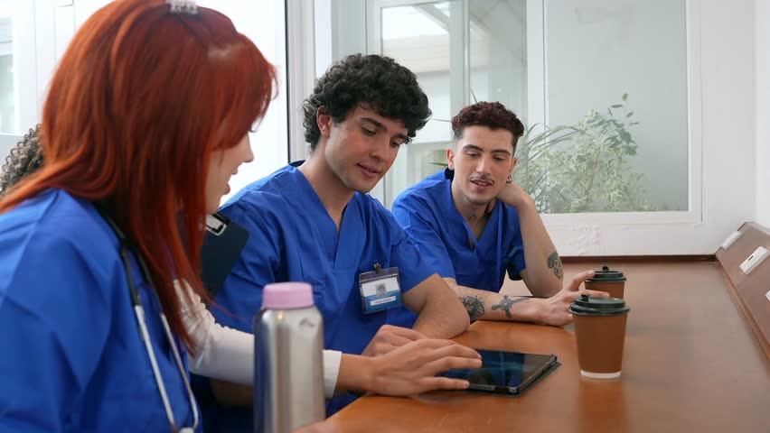 Three young nurses in blue scrubs are sitting together at a table, discussing a case using a digital tablet while drinking coffee during their break in the hospital common area
