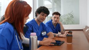 Three young nurses in blue scrubs are sitting together at a table, discussing a case using a digital tablet while drinking coffee during their break in the hospital common area - Powered by Shutterstock - Get 15% off with code: PIKWIZARD15