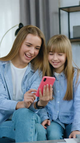 Lovely cheerful trendy younger and eldest sisters sitting one near one on comfortable sofa in beautifully designated living-room and revisioning amusing pictures or photos on mobile