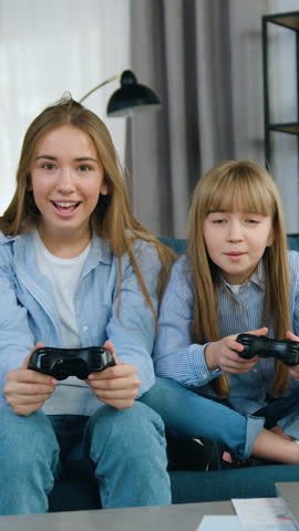 Front view of gorgeous cheerful excited 20-aged and 10-aged girls with blond hairs which enjoying video games applying joysticks when spending joint fre time on soft couch in contemporary apartment
