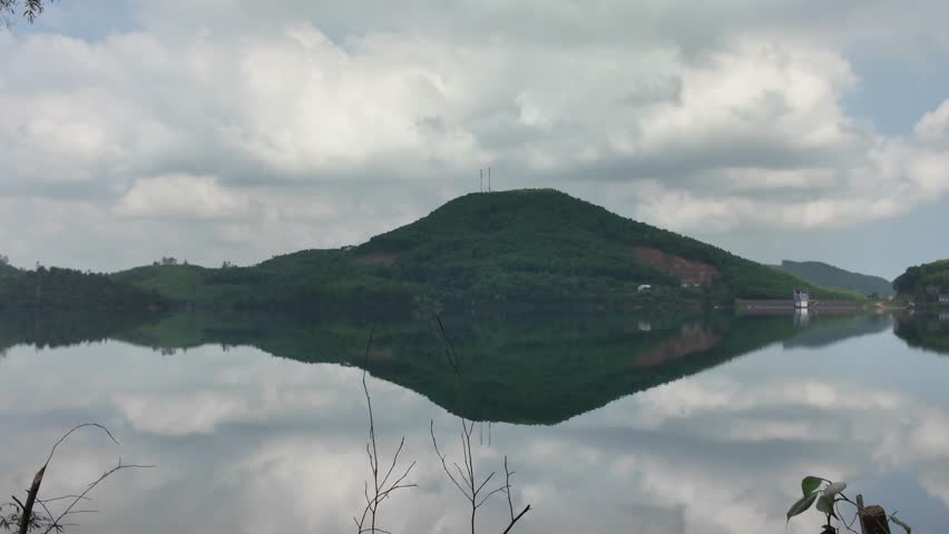 Scenic landscape featuring a green mountain perfectly reflected in the calm, mirror-like surface of a lake under a cloudy sky.