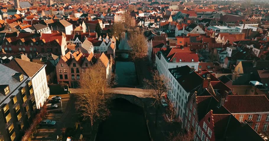 Breathtaking aerial drone footage of the historic canals in Bruges, Belgium. Charming old buildings with red-tiled roofs line the waterway, crossed by a picturesque stone bridge under warm sunlight.
