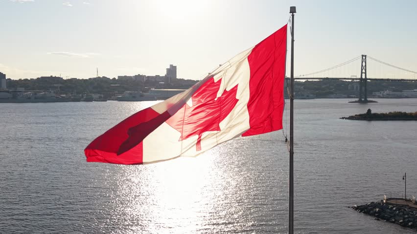 Cinematic drone footage: Canadian flag flutters strongly on Dartmouth shoreline flagpole. Halifax skyline shines in warm summer sunset light, representing national pride and natural beauty 4K.
