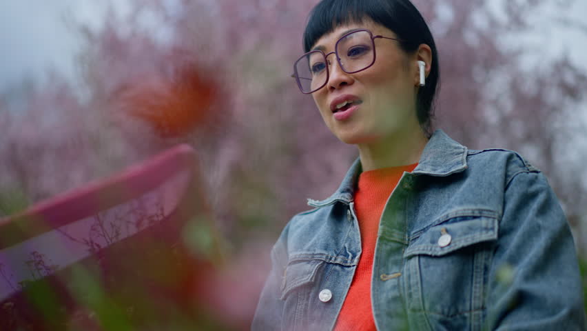 A stylish young woman wearing glasses and wireless earbuds smiles while walking through a park with soft pink blossoms in the background