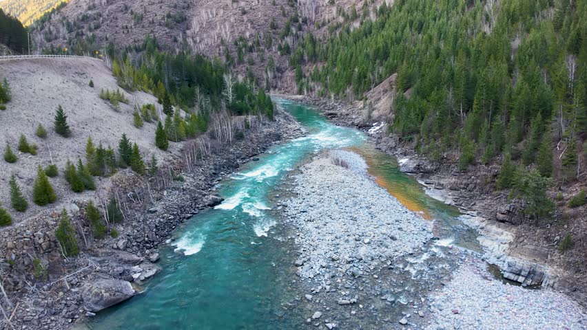 Drone flight along a turquoise river winding through a forested mountain valley and rocky gravel bars in Montana near Glacier National Park.