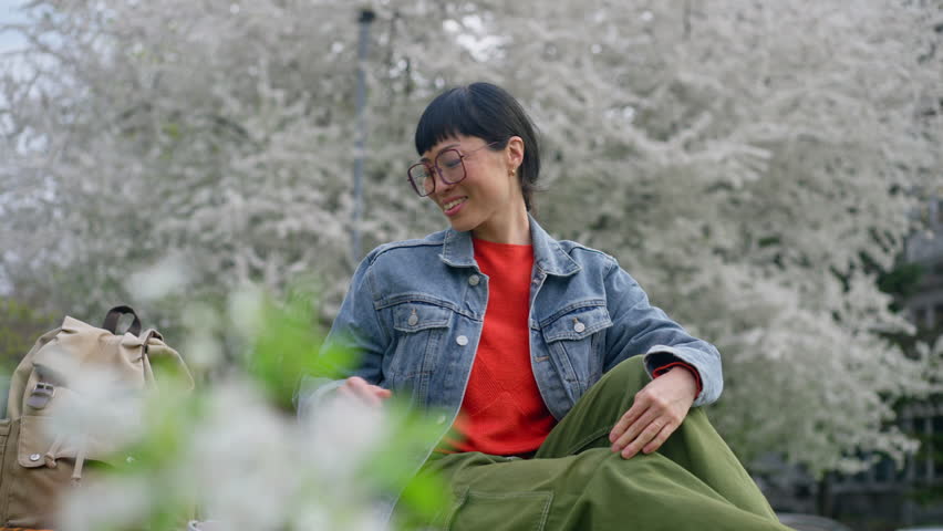 A young woman with headphones on sits outdoors, looking at her phone and smiling amidst blooming trees