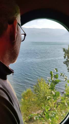 thoughtful man with glasses looking out moving train window, contemplating passing beautiful lake and mountain landscape during his relaxing travel journey on sunny day. vertical.