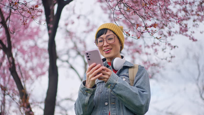 A cheerful young woman wearing glasses and a yellow beanie, with headphones around her neck, engages in a phone conversation while standing under a canopy of pink cherry blossoms
