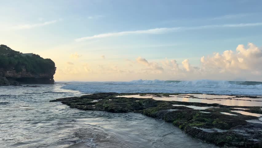 A serene coastal scene with gentle waves washing over rocky shoreline under a soft blue sky at sunrise.