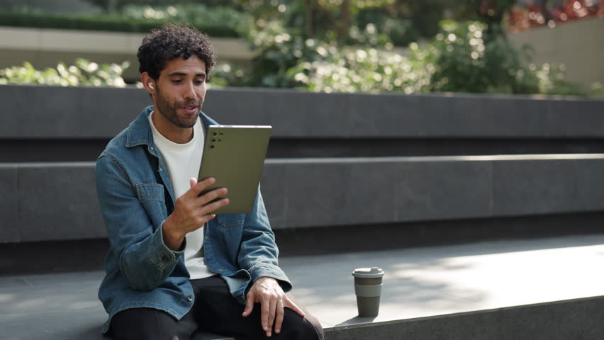 Portrait of curly haired Hispanic man talks on video call via digital tablet in city park. Latin guy discusses college news with friend on online chat via device as sits on parapet
