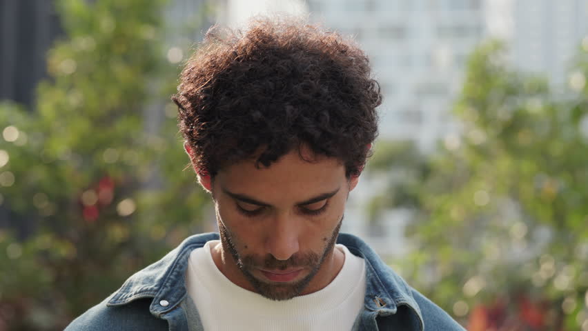 Portrait of serious Hispanic man with curly hair stands in green urban park closeup. Bearded Latin American male with tranquil expression looks at camera on summer city street