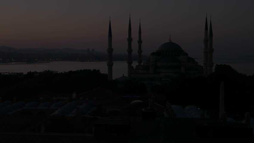 A peaceful sunset scene shows a mosque with tall minarets overlooking a wide body of water and distant city skyline. The soft gradient sky and fading city lights create a calm urban evening atmosphere