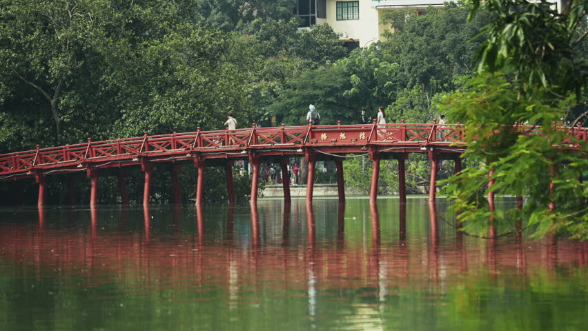 Tourists locals walking across Huc Bridge, prominent traditional red wooden footbridge with intricate design, connecting shore to Ngoc Son Temple Jade Island within Hoan Kiem Lake, Hanoi, Vietnam.