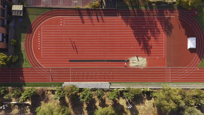 Top-down aerial view of a person running on a bright red track at Colegio Cumbres, featuring a long jump sand pit and surrounding trees. Chile, 4K