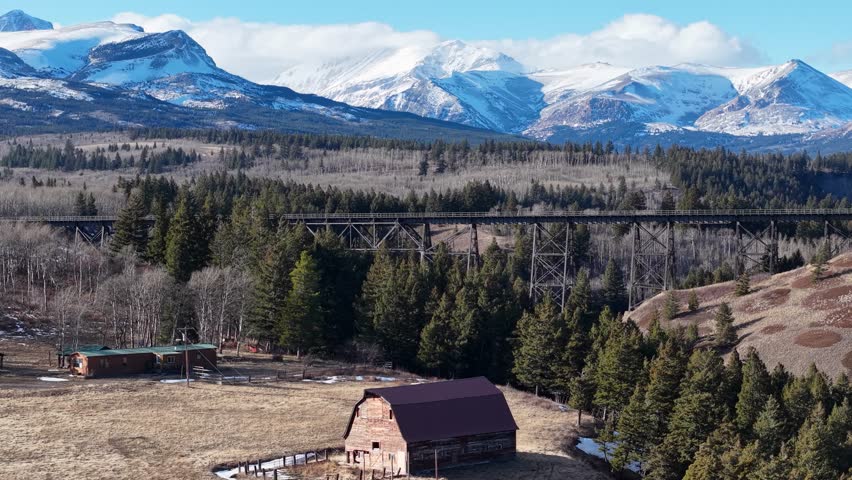 Wide drone shot over a rustic barn and homestead on an open field surrounded by sparse trees with snow-capped mountains rising in the distance near Glacier National Park in Montana.