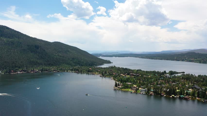 Drone shot over a broad mountain reservoir with sailboats, motorboats, wooded shoreline, and lakeside neighborhoods framed by rolling high country hills in soft daylight