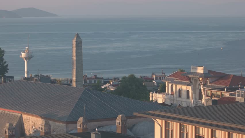 A seaside view in Istanbul shows historic buildings, a stone tower, and calm blue water under soft morning light. The peaceful coastal scene combines architecture and nature in a warm atmosphere.
