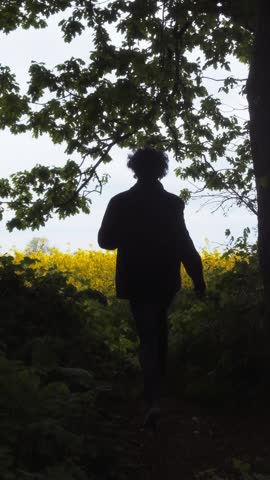 Vertical 1080p back view of a young man running from a dark forest into a vibrant yellow field of rape flowers in bright midday sun. Slow motion with a camera push in.