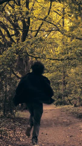 Vertical 1080p slow motion of a man running down a long forest path at midday. Sunlight filters through trees with a slow camera push in from behind.