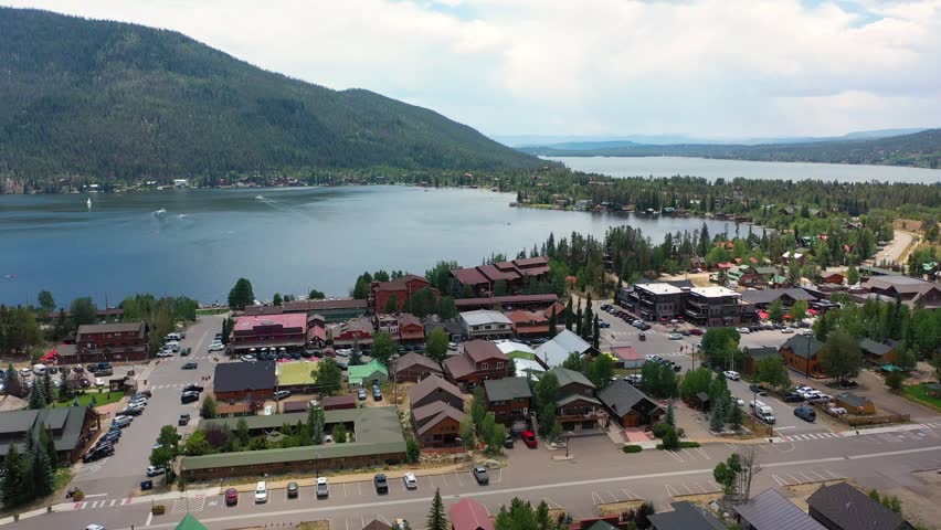 Drone panorama across Grand Lake Colorado showing the lakeside town edge, marina docks, calm mountain water, and evergreen slopes rising toward distant peaks