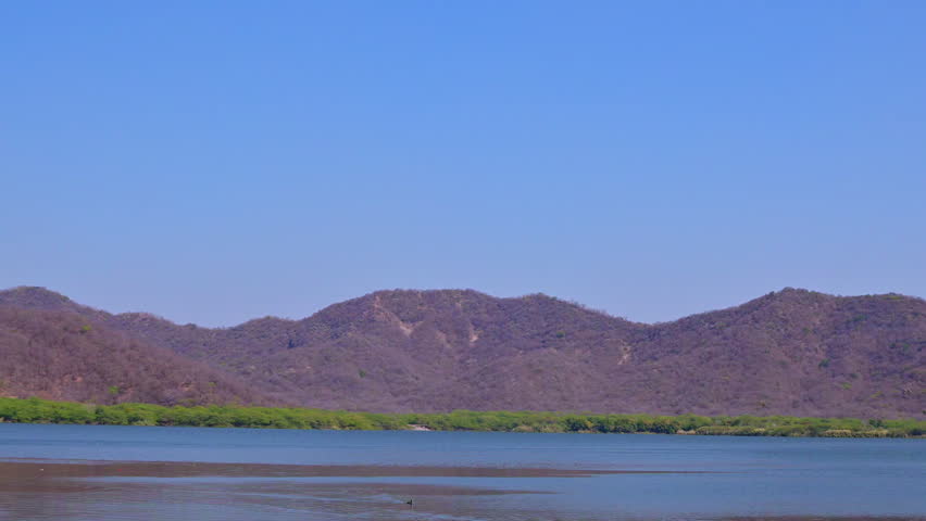 Wide video view of Man Sagar Lake in Jaipur Rajasthan with Aravalli hills in the background, calm water surface in foreground and natural lakeside vegetation visible near Jal Mahal area.