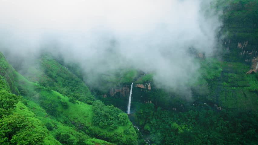 Aerial view of ring waterfall hidden in fog. Beautiful waterfall surrounded by lush green hills and monsoon mist near Tamini Ghat, Maharashtra, India. Clouds covering green valley.
