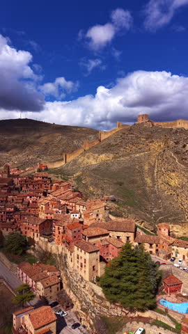 Aerial view of Albarracin with fortified medieval walls climbing rugged hills, terracotta rooftops on a cliffside and dramatic clouds over Aragon landscape