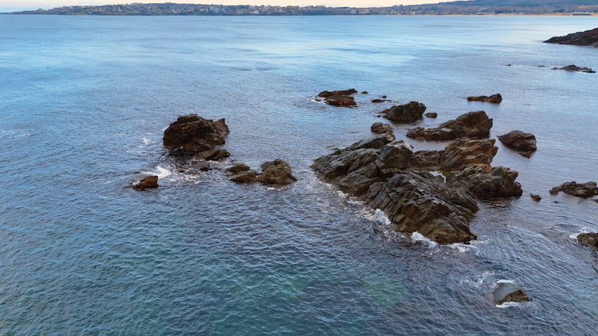 Dark volcanic rocks in calm blue sea water. Group of jagged dark stones protruding from the ocean surface with a town in the distance.