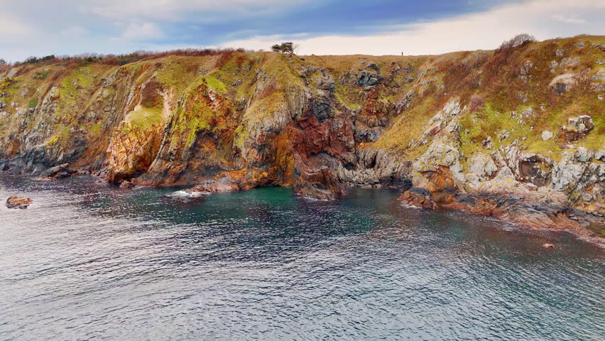 Close up of layered rock formations by sea water. Detail shot of textured geological cliffs with red and brown minerals near blue water.
