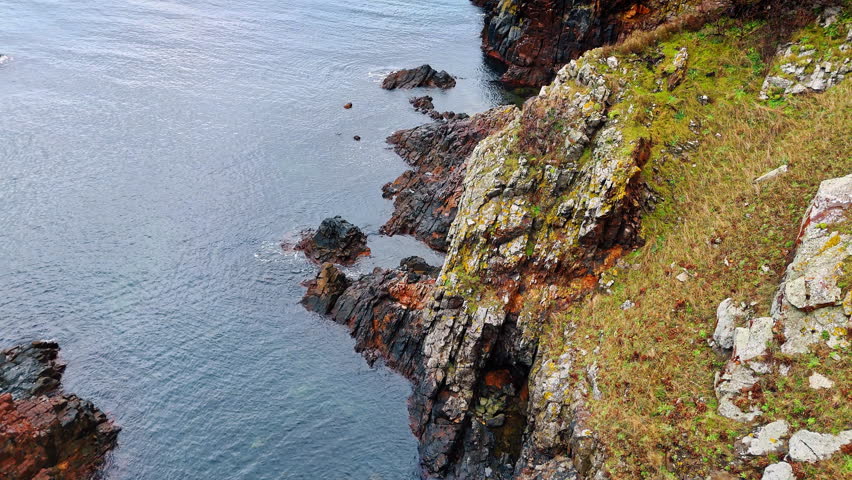 High angle view of rocky cliff edge and sea. Vertical perspective of a steep grassy cliff side descending into dark ocean water.