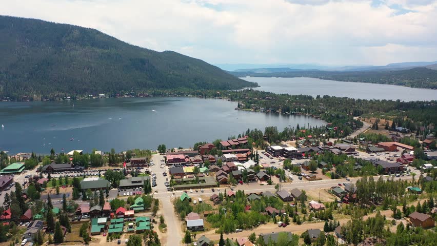 Aerial panorama of Grand Lake Colorado with lakeside buildings, marina piers, forested slopes, and calm water backed by rugged Rocky Mountain scenery in summer