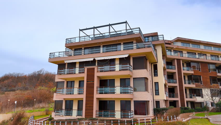 Modern multi story apartment building with balconies near the forest. Front view of a contemporary residential complex with glass railings and wood panels under a cloudy sky.