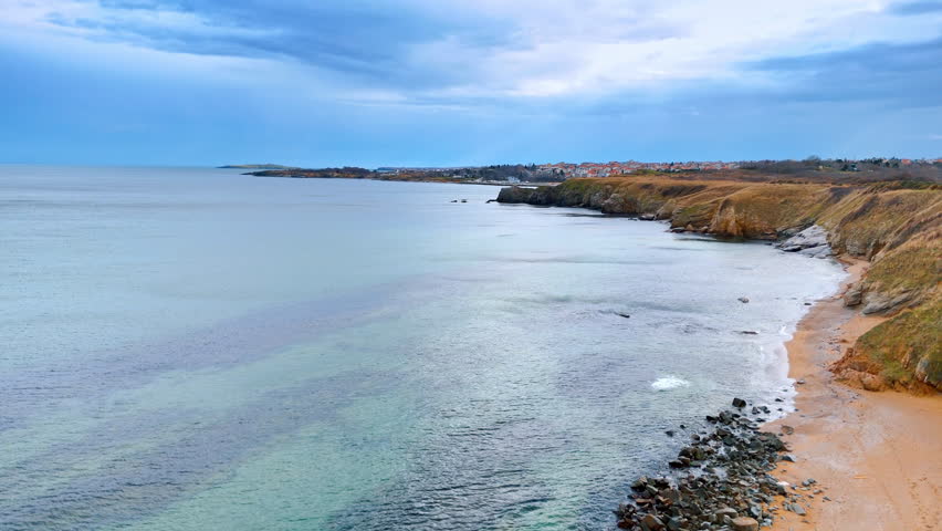 Coastal landscape with sandy beach and rocky cliffs under cloudy sky. Wide angle view of a curved coastline featuring a sandy beach, rocky outcrops, and distant coastal village.