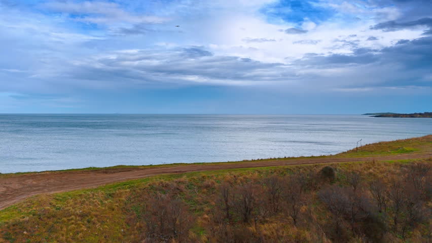 Wide view of sea horizon from a coastal dirt road. Panoramic landscape showing a dirt path along a grassy cliff with a calm sea under a cloudy sky.