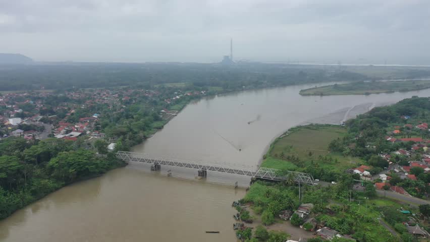 River flows wide under cloudy skies. Bridge spans water, connecting towns and green fields. Fishing boats drift peacefully along the current. Village rooftops peek through lush trees