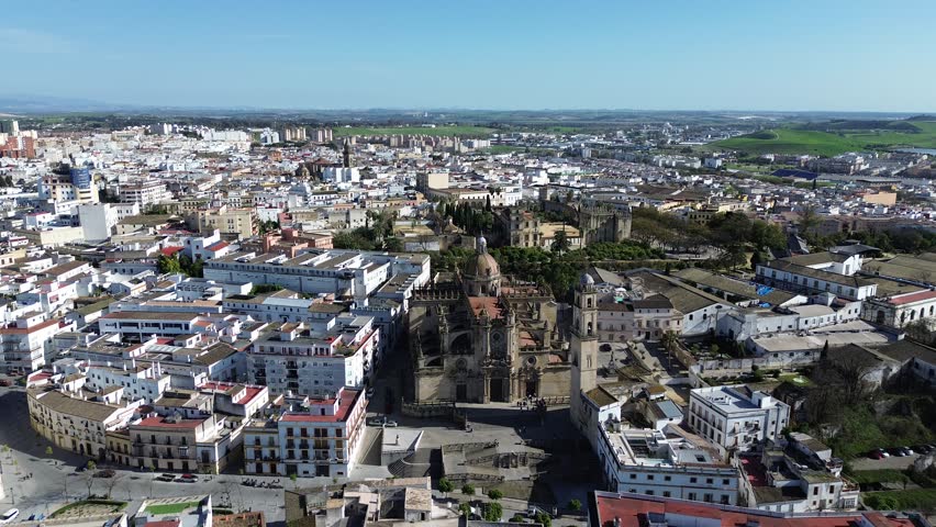 Aerial drone shot moving forward in 4K resolution at high altitude approaching the main facade of the Jerez Cathedral with the historic Alcazar fortress visible behind it under a clear sunny sky