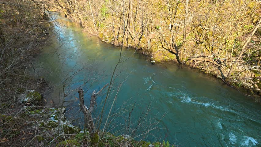 High angle view of a turquoise river flowing upstream, filmed from a tall rock or cliff. The camera follows the river’s course, capturing natural water movement and surrounding autumn forest on a sunny day with clear sky. Ideal for cinematic, travel, and nature projects.