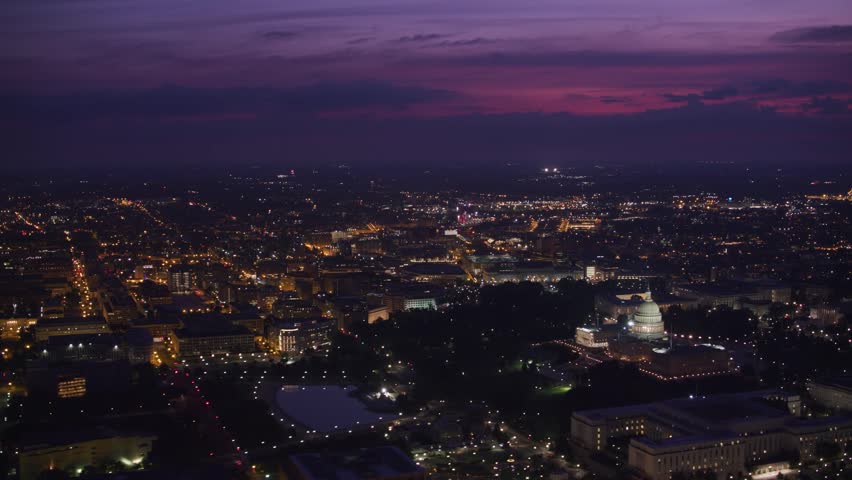 Sweeping aerial drone shot of the Washington D.C. skyline at dusk. The city is illuminated with glowing streetlights under a dramatic purple and pink twilight sky, featuring the iconic United States Capitol building brightly lit in the distance.